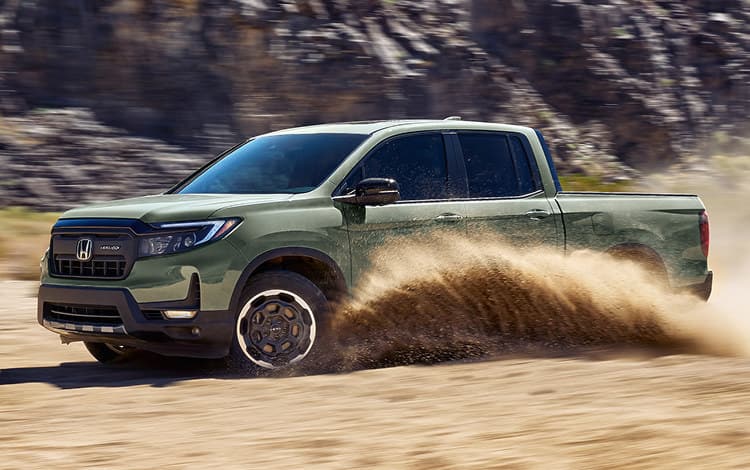 2026 Ridgeline TrailSport in Ash Green Metallic driving  through rugged terrain with dust kicking up,  with mountains in background, at sunrise.