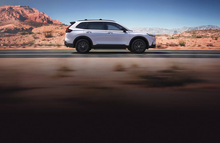 2027 Honda CR-V Sport Touring Hybrid in Platinum White Pearl profile driving on a scenic desert road (Compact Crossover SUV).