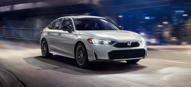2027 Honda Civic Sedan Sport Hybrid in Solar Silver Metallic front driving on a city street at night (Compact Sedan).